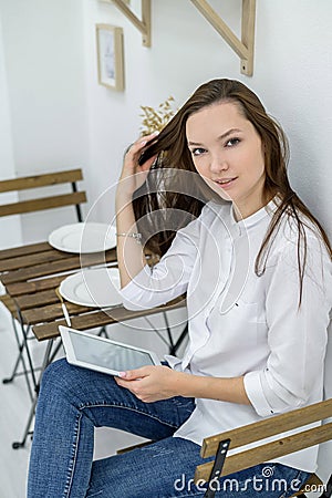 A young woman in a white shirt and jeans sits in a cafe at a table with a tablet in her hands. Female office worker at Stock Photo