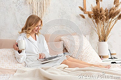 Young woman in white terry robe is drinking coffee and reading magazine or book in bedroom. Stock Photo