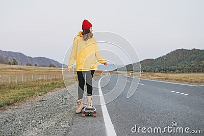 Young woman in yellow hoodie and red hat on skateboard on the road against beautiful mountain landscape, Chuysky tract, Altai Stock Photo