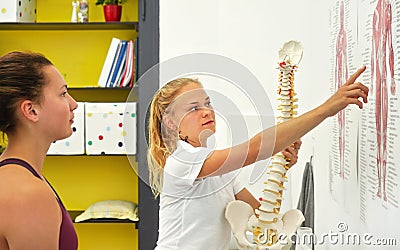 Young woman patient at physiotherapy consultation, physiotherapist explaining back bones on human muscles body diagram, holding Stock Photo