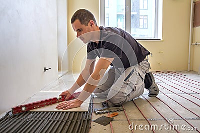 Young worker tiler installing ceramic tiles using lever on cement floor with heating red electrical cable wire system. Home Stock Photo