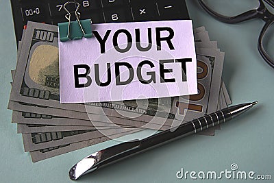 YOUR BUDGET - words on a white piece of paper fixed on banknotes against the background of a calculator, glasses and pen Stock Photo