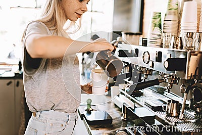 A youthful slim kind blonde girl,dressed in casual outfit,skillfully adds milk to coffee in a modern coffee shop. Stock Photo