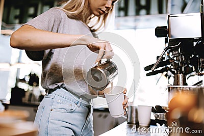 A youthful thin blonde girl,wearing casual cothes,is shown adding milk to the coffee in a cozy coffee shop. Stock Photo