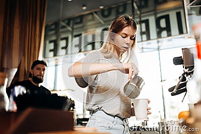 A youthful thin blonde girl,wearing casual cothes,is shown adding milk to the coffee in a cozy coffee shop. Stock Photo