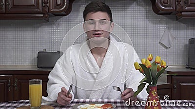 Привлекательный молодой человек в белом купальном халате сидя на кухонном столе наслаждаясь ароматностью плиты завтрака с зажарен акции видеоматериалы Привлекательный молодой человек в белом купальном халате сидя на кухонном столе наслаждаясь ароматностью плиты завтрака с зажарен акции видеоматериалы