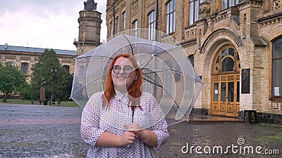 Тучная девушка имбиря с стеклами стоит в парке в ненастной погоде, держащ зонтик, наблюдающ на камере, усмехаясь Тучная девушка имбиря с стеклами стоит в парке в ненастной погоде, держащ зонтик, наблюдающ на камере, усмехаясь