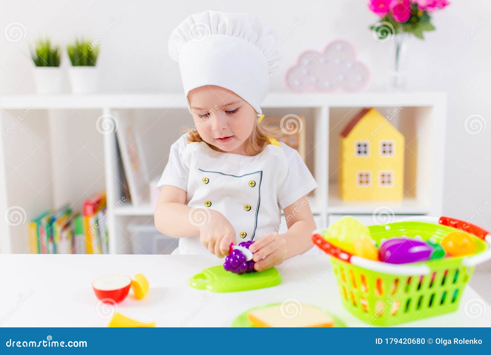 μικρή ξανθιά κοπέλα με λευκή στολή μάγειρας που παίζει με παιδικά φρούτα  και λαχανικά στο σπίτι στο νηπιαγωγείο ή στο νηπιαγωγείο. Στοκ Εικόνες -  εικόνα από childhood, childcare: 179420680