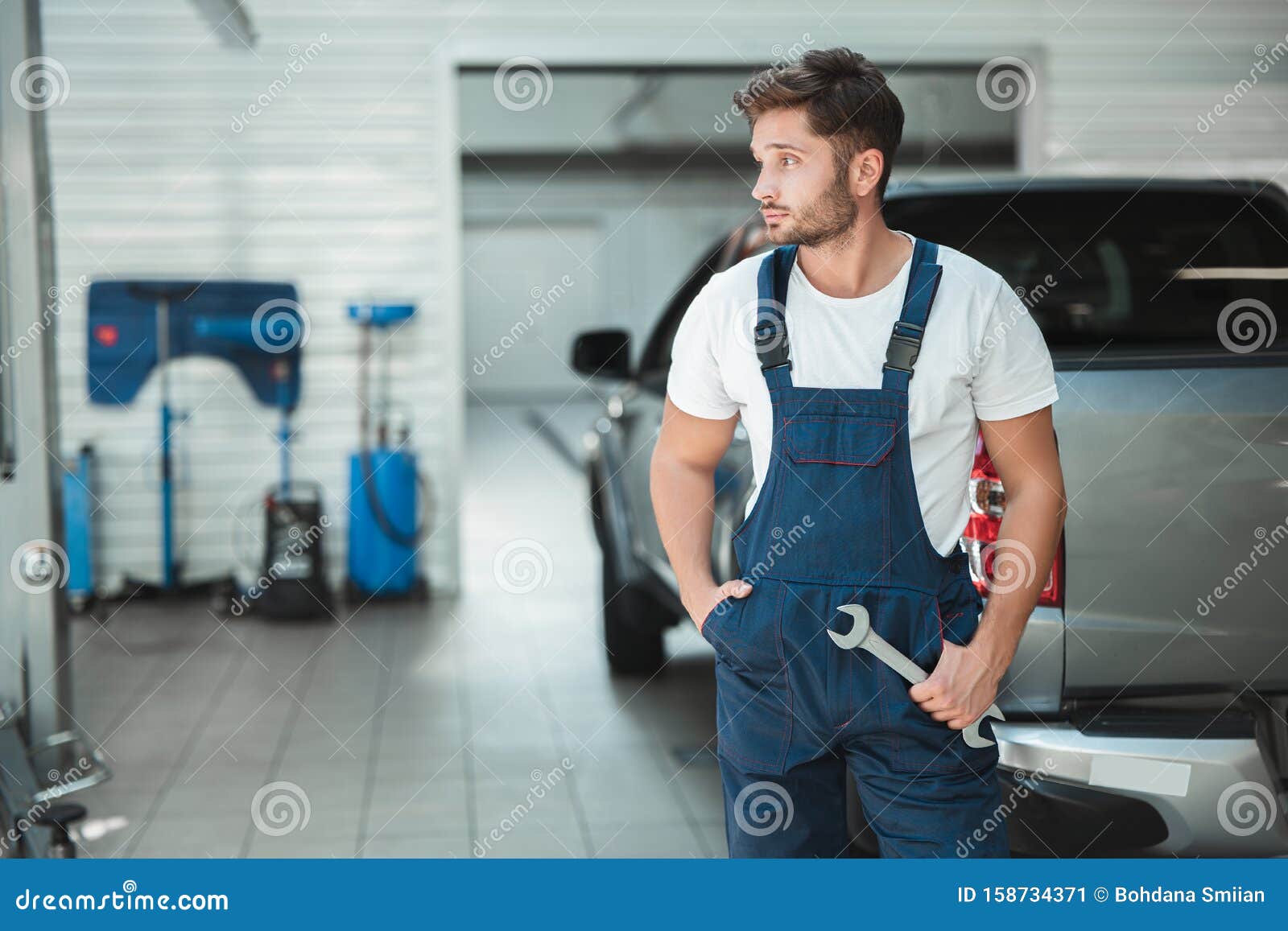 Νεαρός σέξι μηχανικός που φοράει στολή με το ένα χέρι στην τσέπη και με ένα  χέρι σε ένα άλλο πόδι στέκεται στο αυτοκίνητο Στοκ Εικόνα - εικόνα από :  158734371