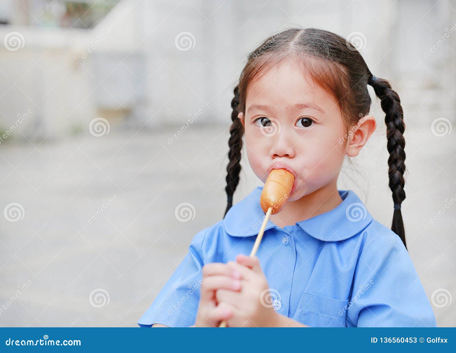 Το πορτρέτο λίγου ασιατικού κοριτσιού παιδιών στη σχολική στολή απολαμβάνει  το νόστιμο λουκάνικο Στοκ Εικόνα - εικόνα από childhood, babylonia:  136560453