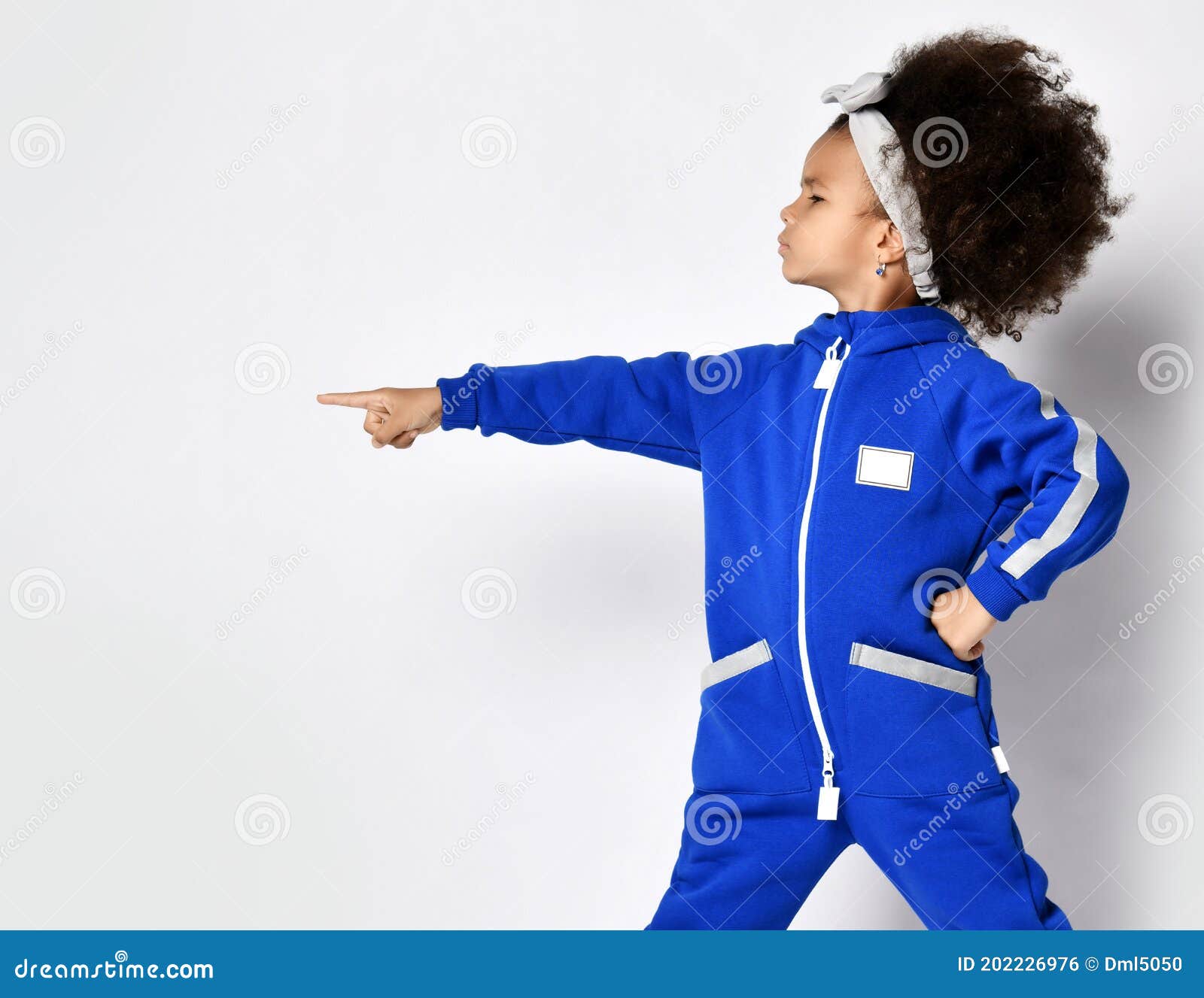 χαριτωμένη κοπέλα με κατσαρό μουστάκι αφρικανή με αναπαυτική μπλε αθλητική  στολή και κορδέλα μαλλιών που στέκονται και δείχνουν στ Στοκ Εικόνες -  εικόνα από lifestyle, childhood: 202226976