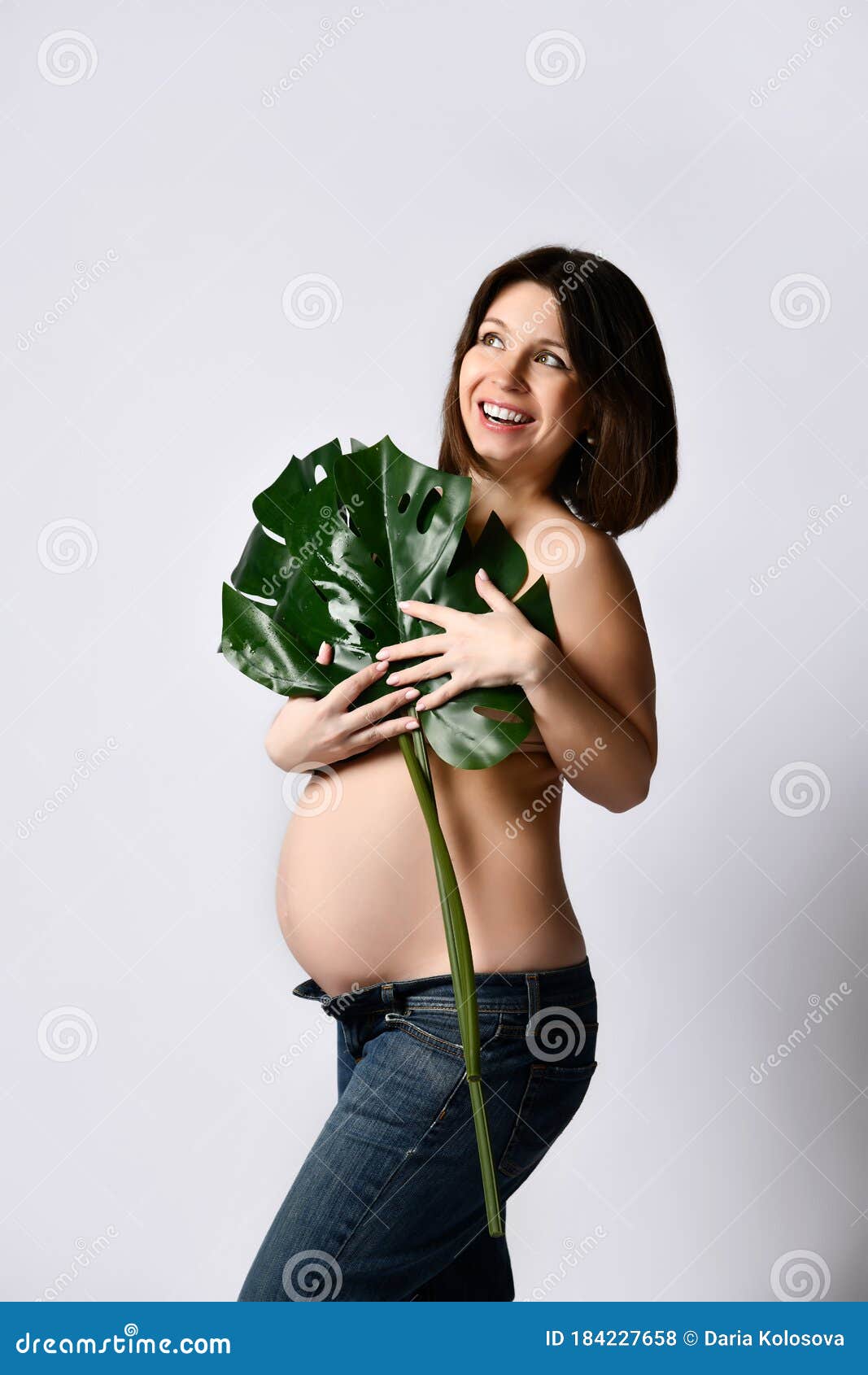 Беременная женщина в джинсах, голые по пояс. Улыбающийся, прикрывая грудь зелеными лист, касающиеся животик, представляющих боком Стоковое Фото - изображение насчитывающей здорово, представлять: 184227658