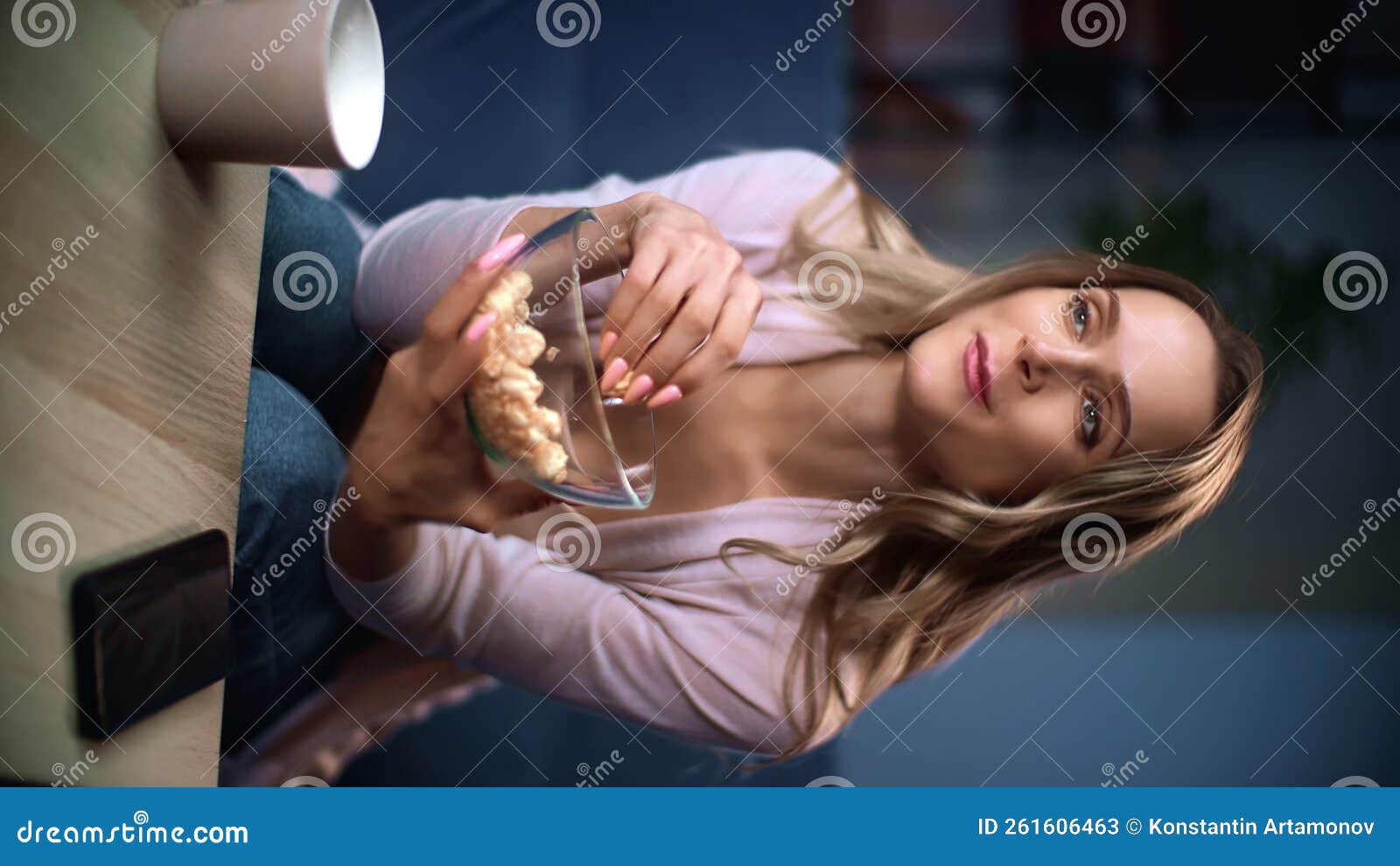 Вертикальное видео, улыбающаяся блондинка, смотрящая телешоу, поедающая попкорн, расслабляющая дома сток-видео - Видео насчитывающей еда, попкорн: 261606463