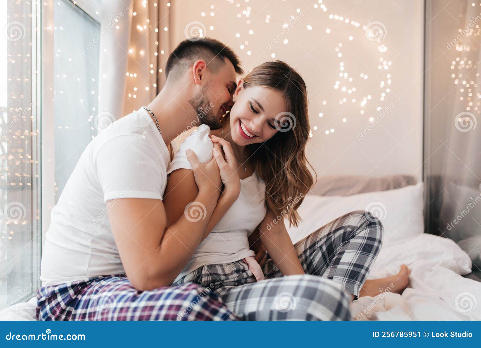 кавказский мужчина в клетчатой пижаме целует свою жену. улыбающаясяблондинка в ночном белье, сидящая на кровати с парнем. Стоковое Изображение- изображение насчитывающей пары, смешно: 256785951
