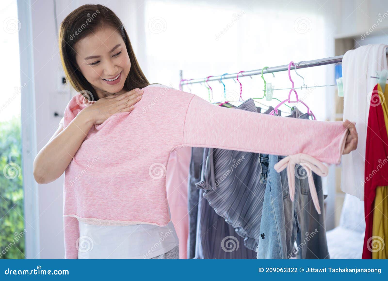 漂亮的亚洲女性在家里的镜门衣橱前试一件粉色毛衣库存照片. 图片  image