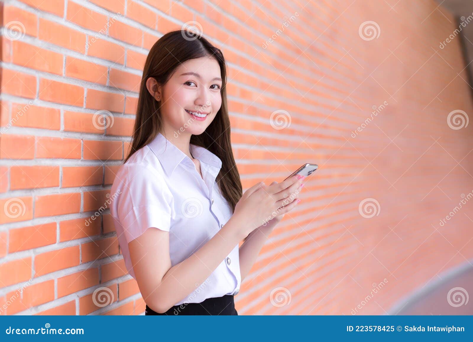身穿制服的亚洲泰国女学生的肖像画在大学里用砖墙的智能手机时高兴地笑  image