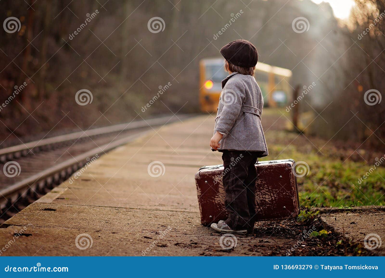 Aanbiddelijke jongen op een station, die op de trein met koffer en teddybeer wachten. De aanbiddelijke jongen op een station, die op de trein met koffer en teddybeer, wijnoogst wachten kijkt