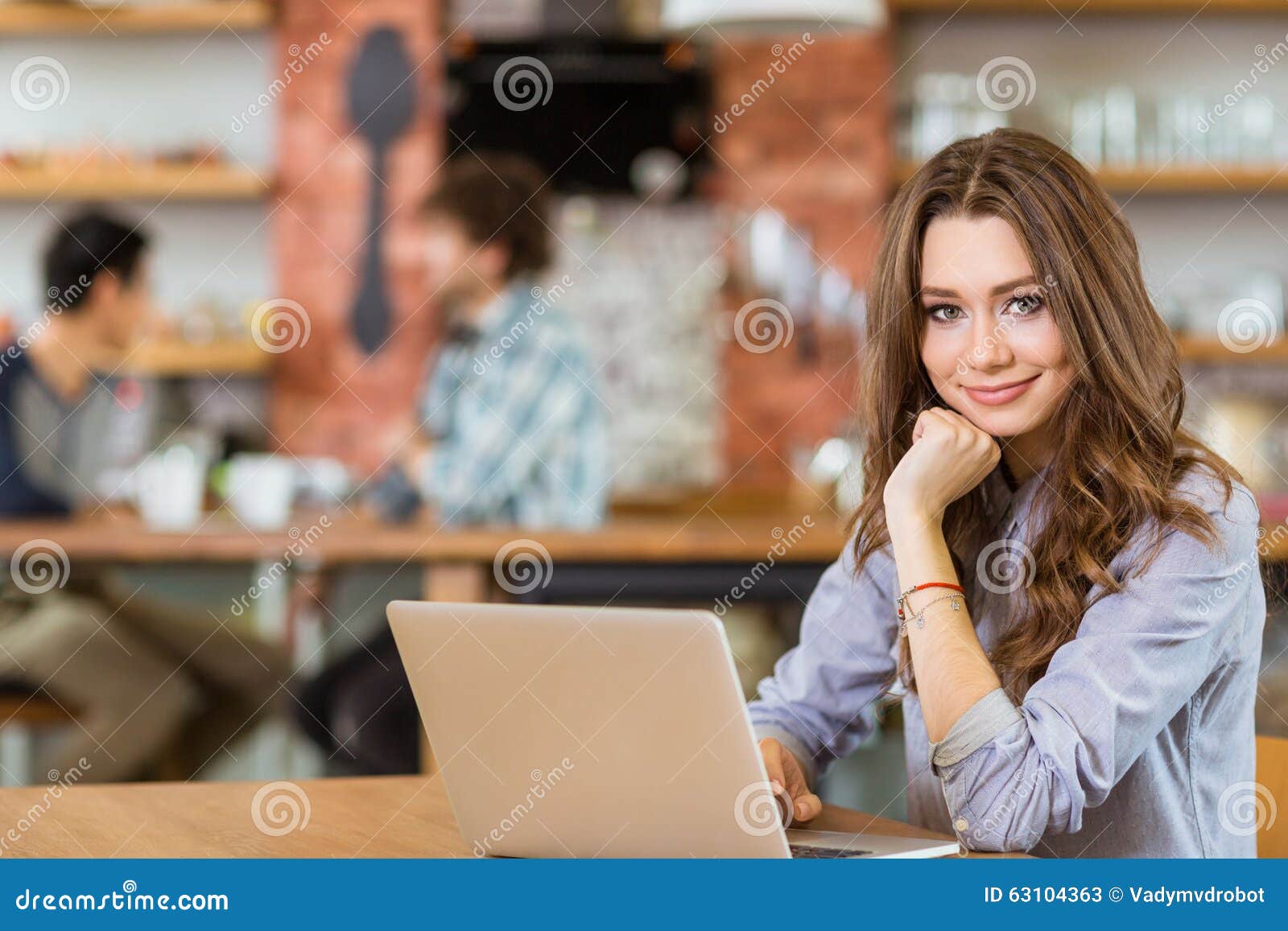 Aantrekkelijke positieve jonge krullende vrouwelijke gebruikende laptop in koffie. Aantrekkelijk positief jong krullend wijfje in grijs overhemd die laptop in koffie met behulp van