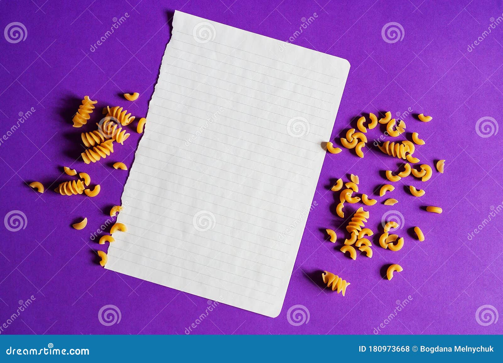 abstraction, on purple background, wicker round stand and on it lies empty piece of paper for text, around scattered colored pasta