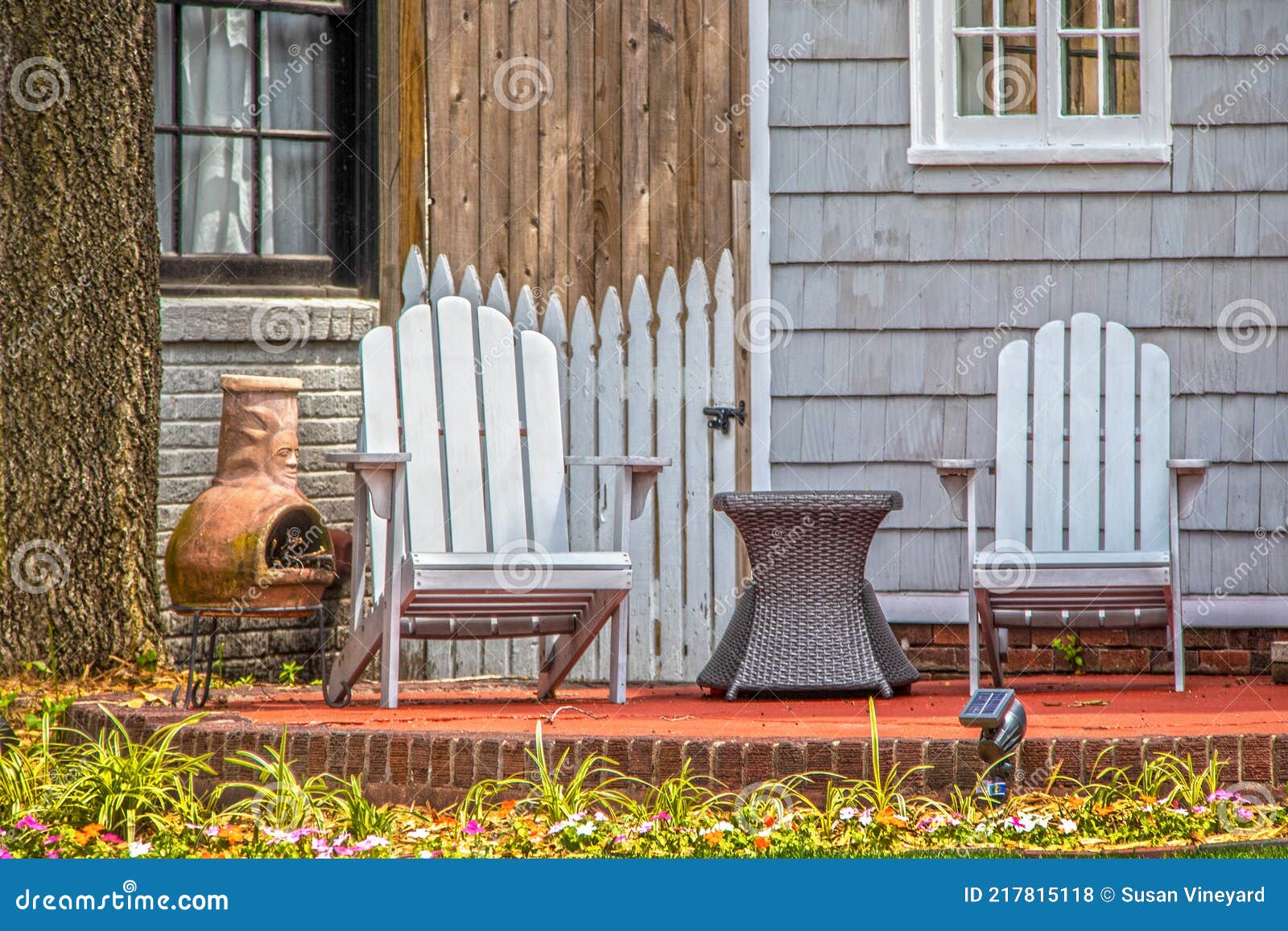 adirondack chairs and chiminea and wicker table on patio under stree in front of house with grey wooden siding and picket fence