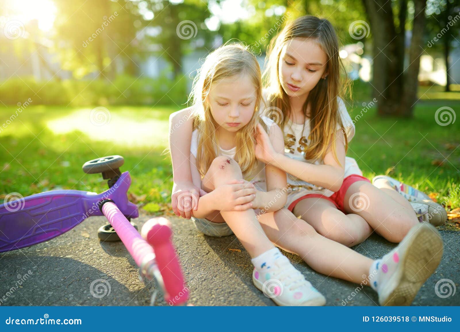 adorable girl comforting her little sister after she fell off her scooter at summer park. child getting hurt while riding a kick s