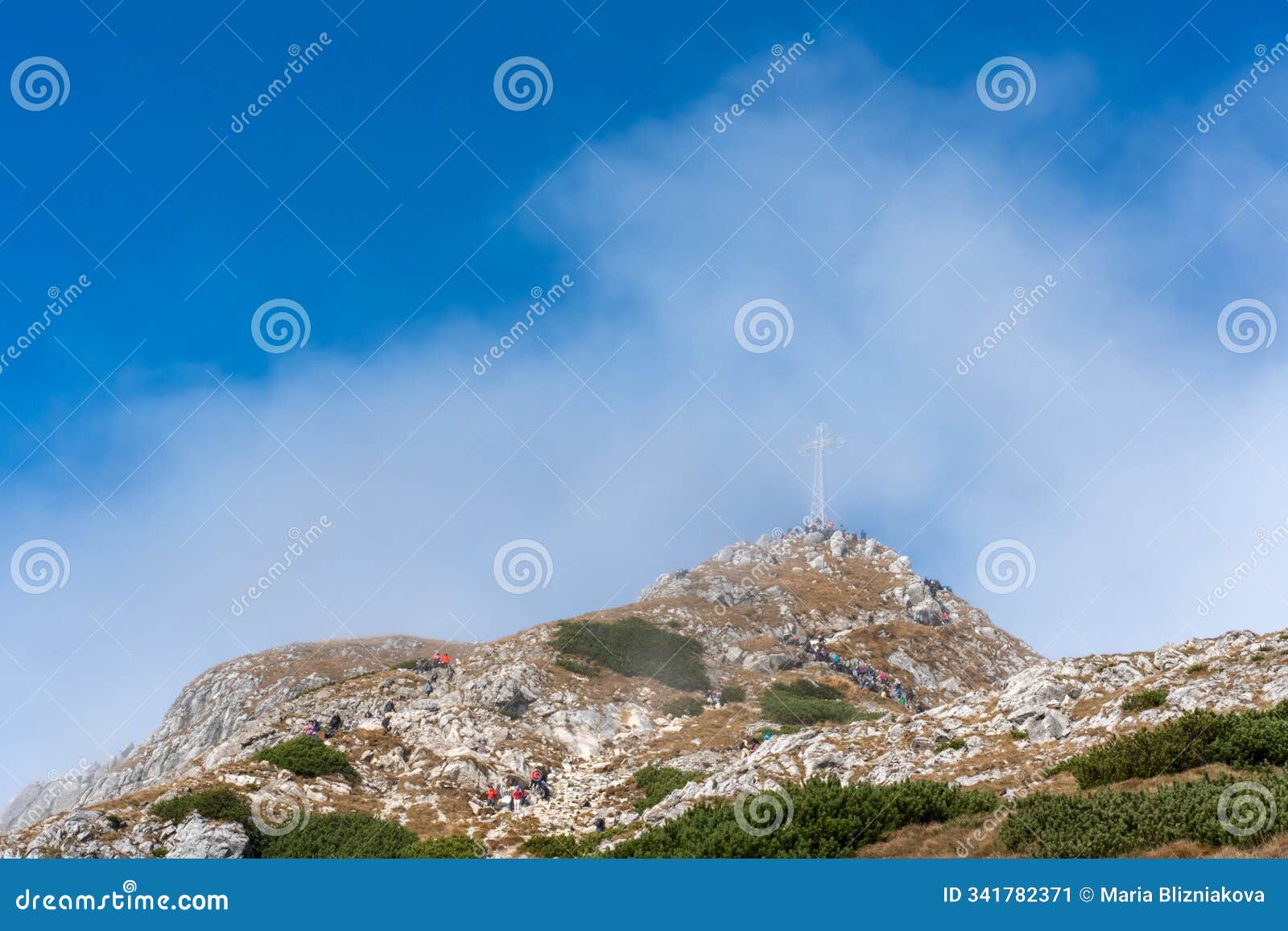 adventurous hikers making their way up a rugged mountain path towards a summit cross on a clear, sunny day.