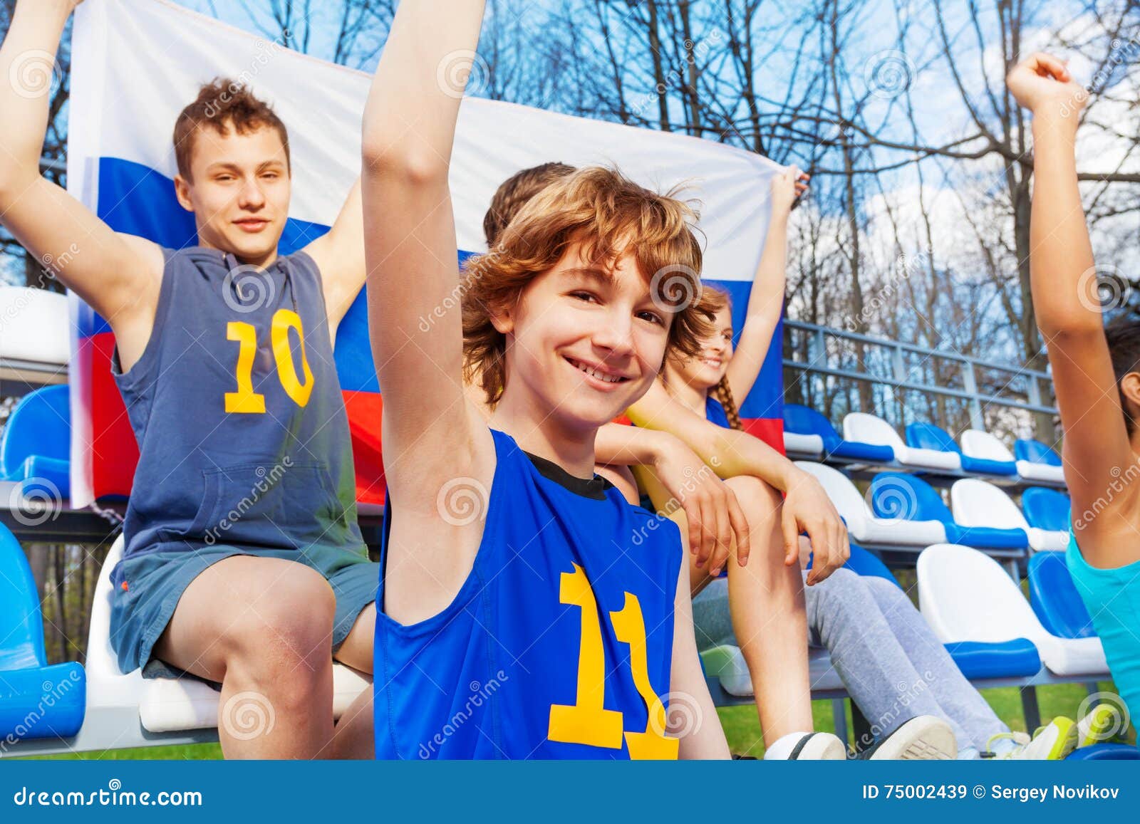 Aficionados deportivos adolescentes felices con la bandera rusa. Retrato del primer de aficionados deportivos adolescentes sonrientes, apoyando a su equipo, sentándose en la tribuna y sosteniendo la bandera de Rusia detrás