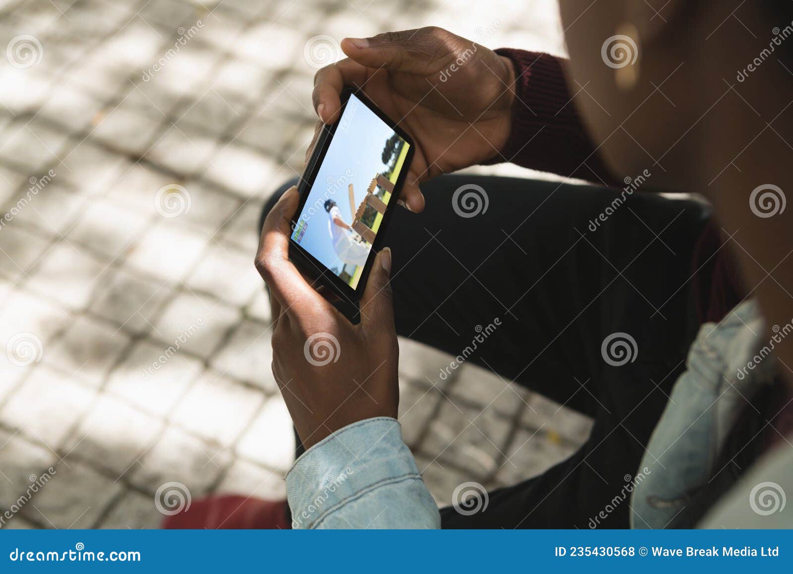 African American Man in Park Watching Cricket Match on Smartphone Stock Photo