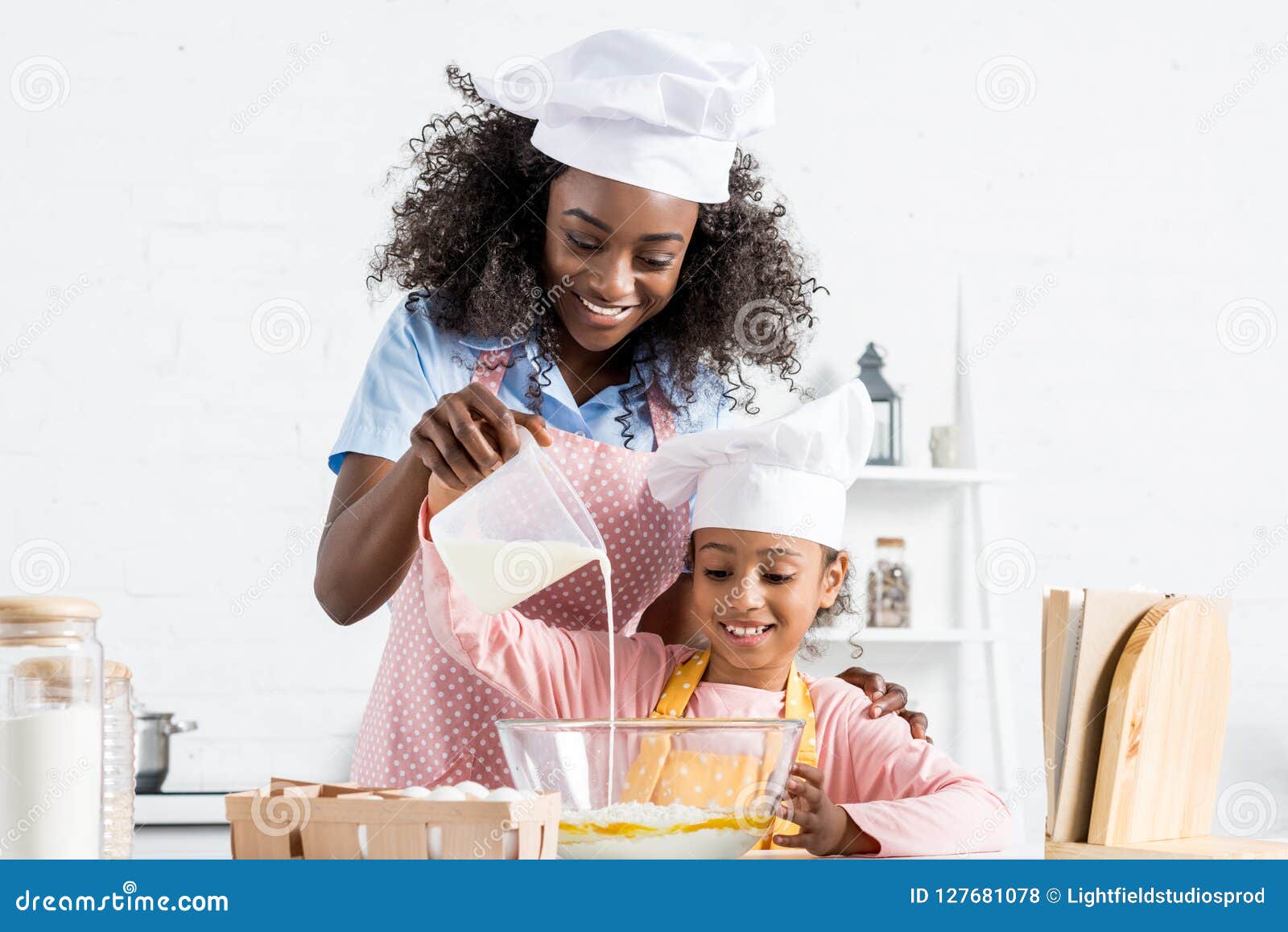 african american mother and daughter in chef hats pouring milk in bowl with ingredientes