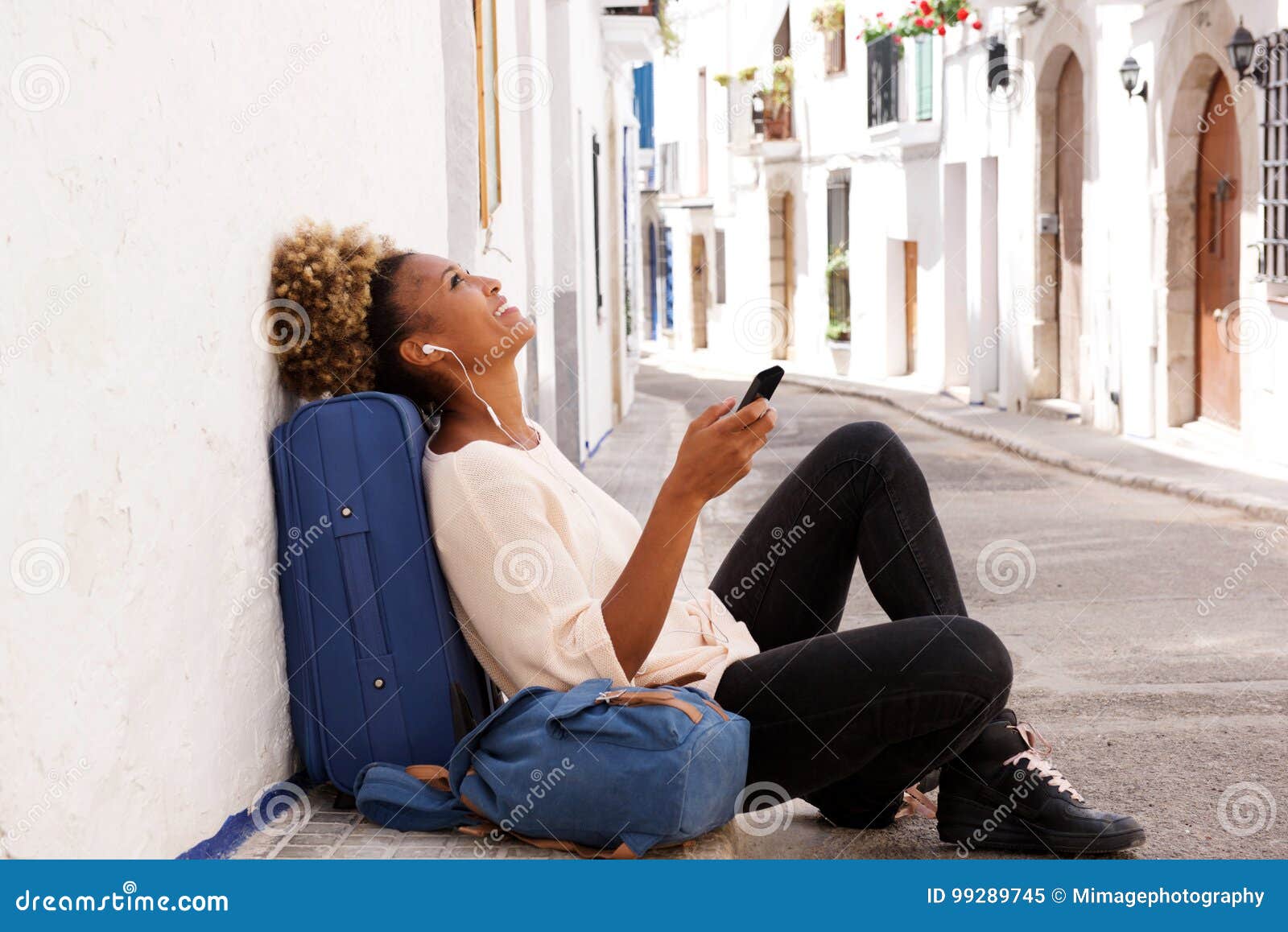 african american traveler female sitting on sidewalk and listening music from smart phone