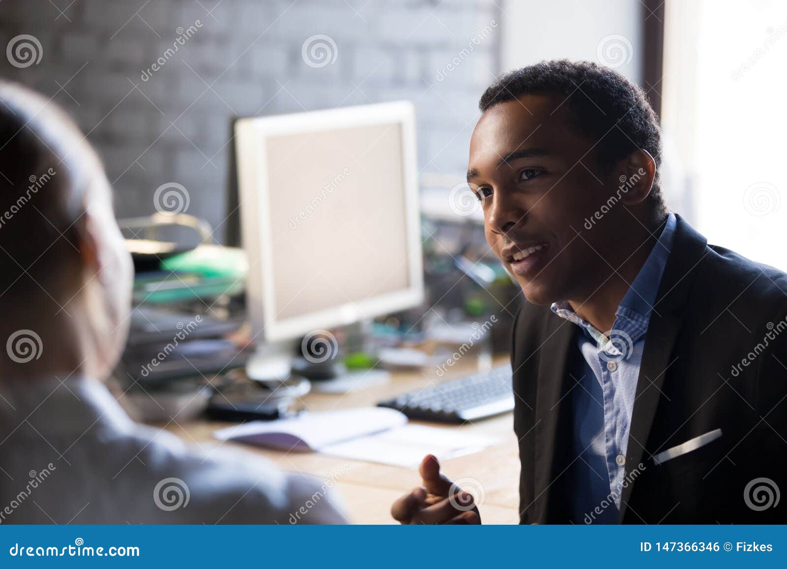 african employee talking with female colleague sitting together at workplace