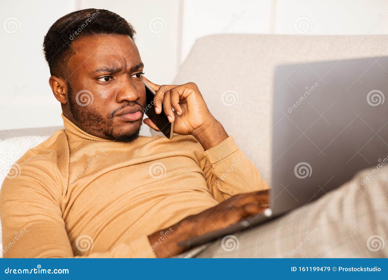 afro guy talking on phone using laptop lying on sofa at home