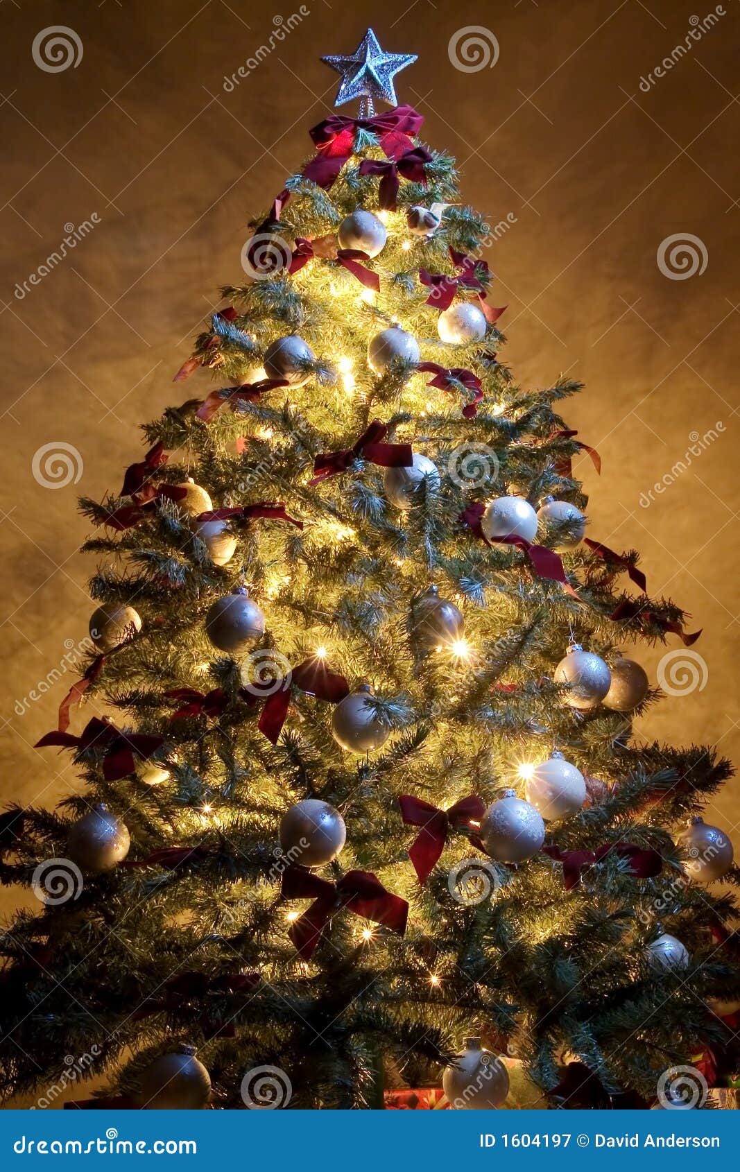 Albero di Natale 3. Un albero di Natale illuminato dall'interno di e circondato dai regali. Decorato con le lampadine bianche ed i nastri rossi. La parete strutturata dietro è illuminata dall'albero.