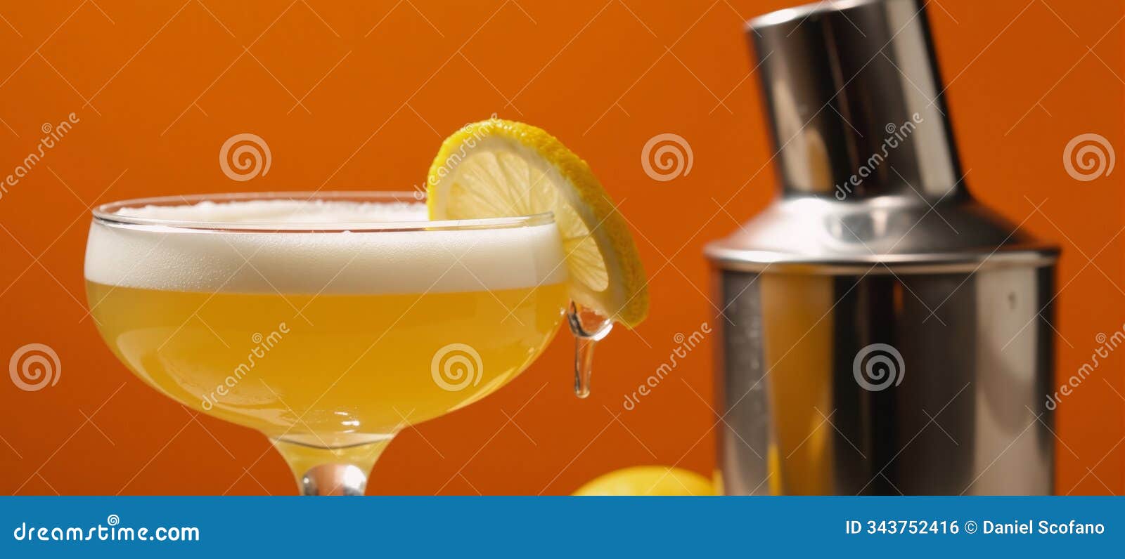 an ambercolored amaretto sour on an orange backdrop with frothy top cocktail shaker and lemon wheel
