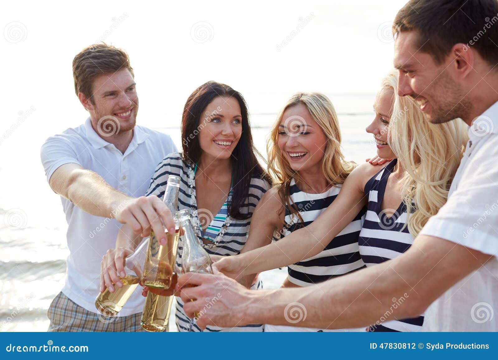 Amigos sonrientes que tintinean las botellas en la playa. Verano, días de fiesta, turismo, bebidas y concepto de la gente - grupo de amigos sonrientes que tintinean las botellas de cerveza o de sidra en la playa