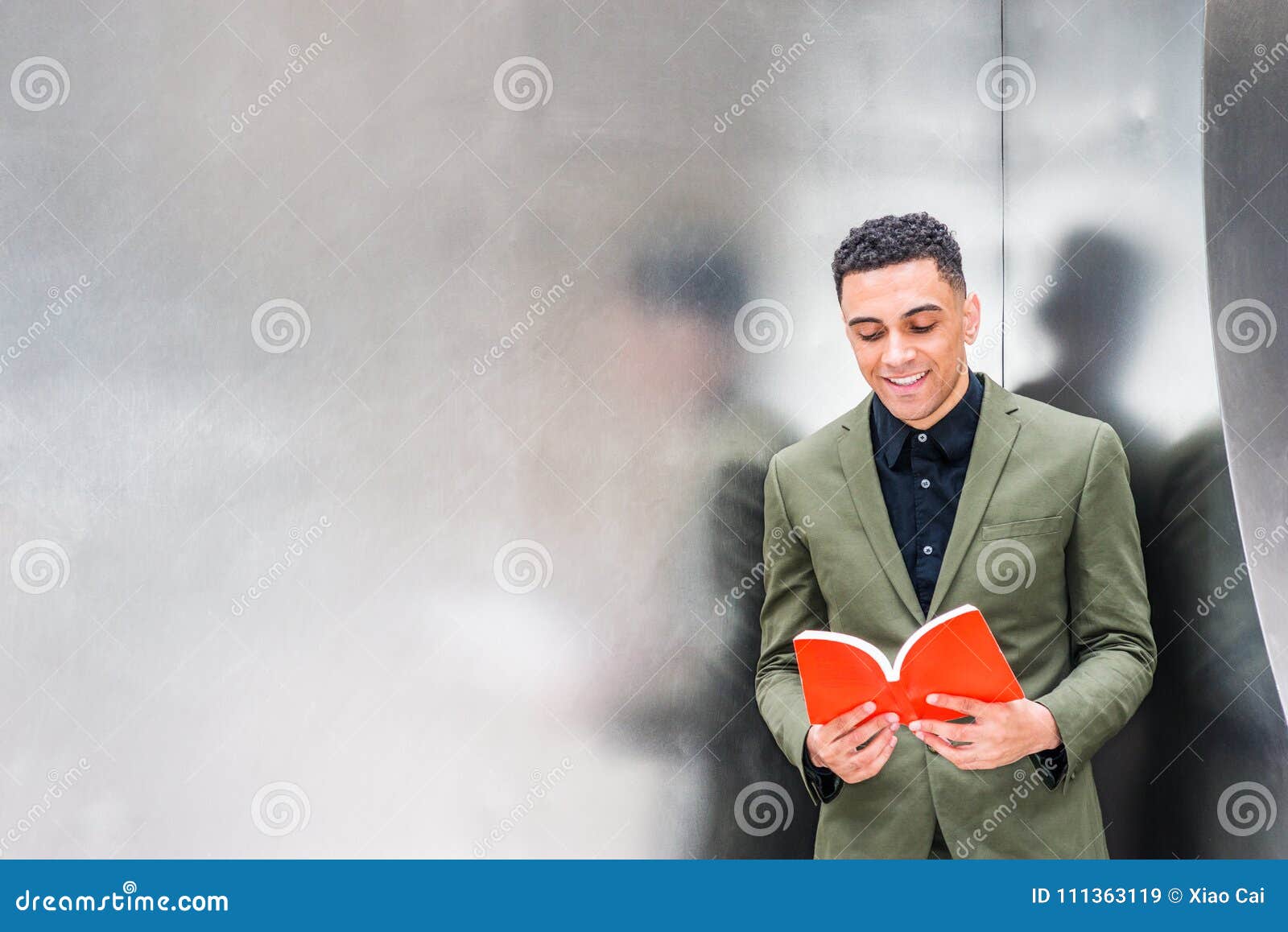 Amo il libro di lettura Giovane uomo d'affari americano bello che indossa vestito verde, stante contro la parete dell'argento del metallo con le riflessioni a New York, guardando giù, leggendo libro rosso, sorridente