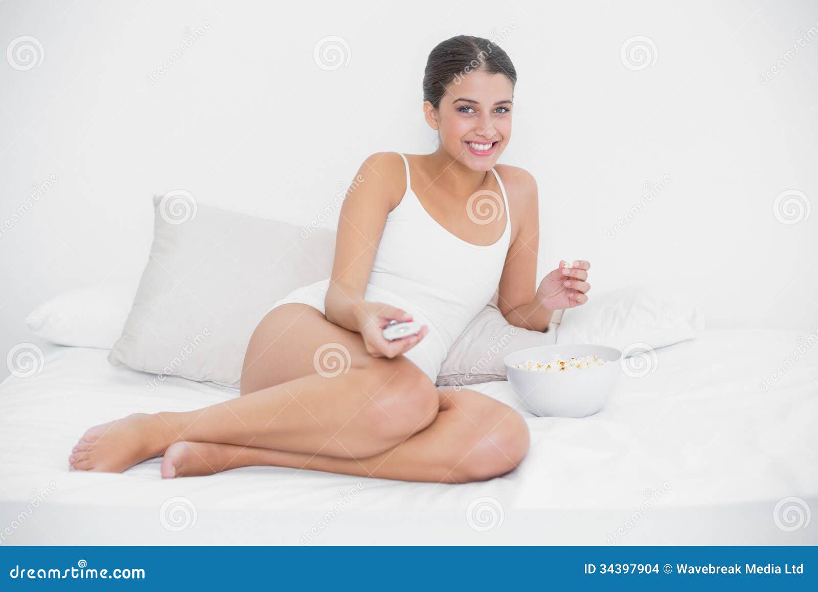 Amused Young Brown Haired Model  In White Pajamas Watching  Tv  While Eating Popcorn Stock Photo 