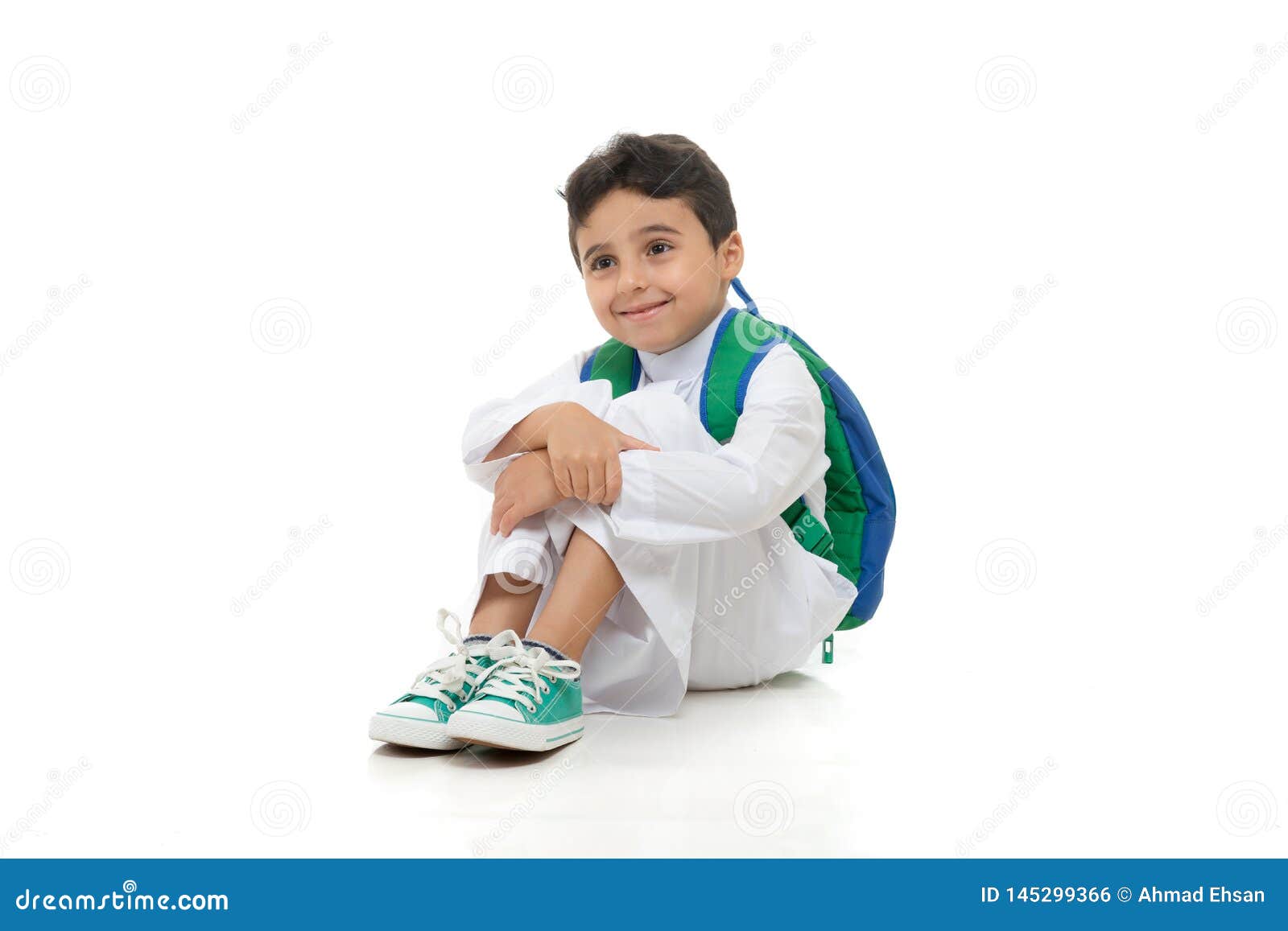 arab school boy sitting on ground with a smile on his face, wearing white traditional saudi thobe, back pack and sneakers, raising