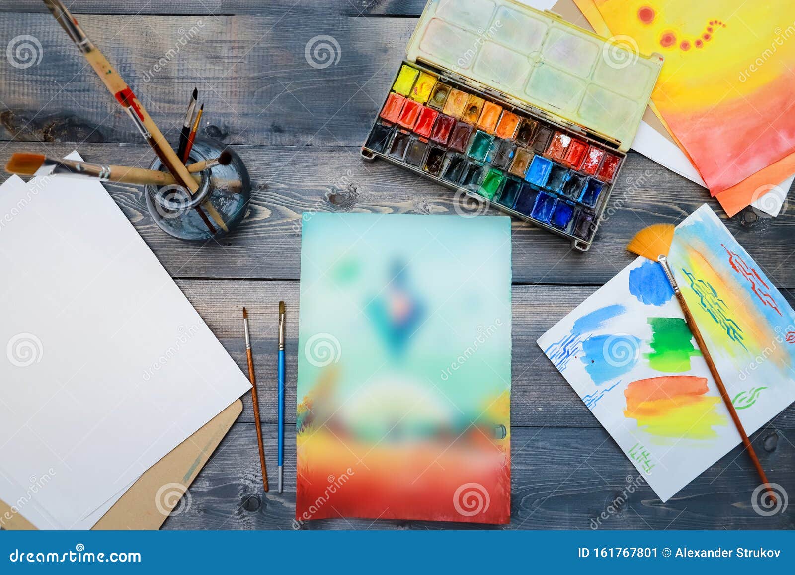 the artistÃ¢â¬â¢s supplies and his hand-made watercolor are laid out on a gray wooden table. on the table are open watercolor paints