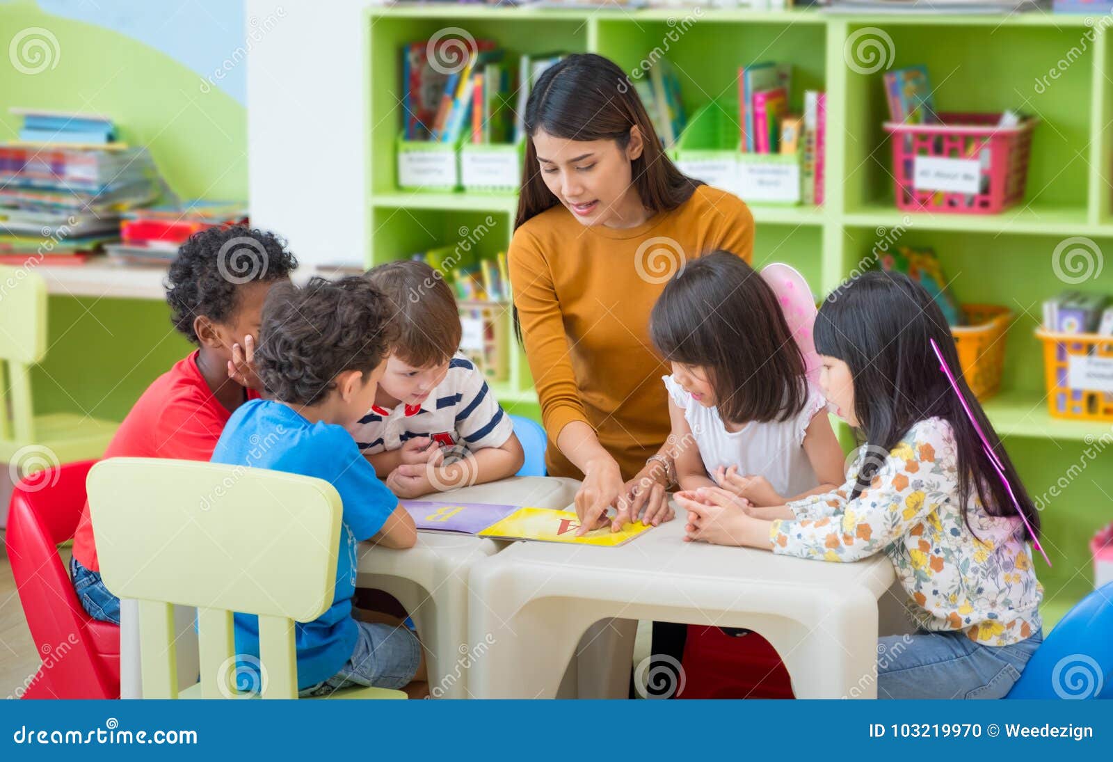 asian female teacher teaching mixed race kids reading book in cl