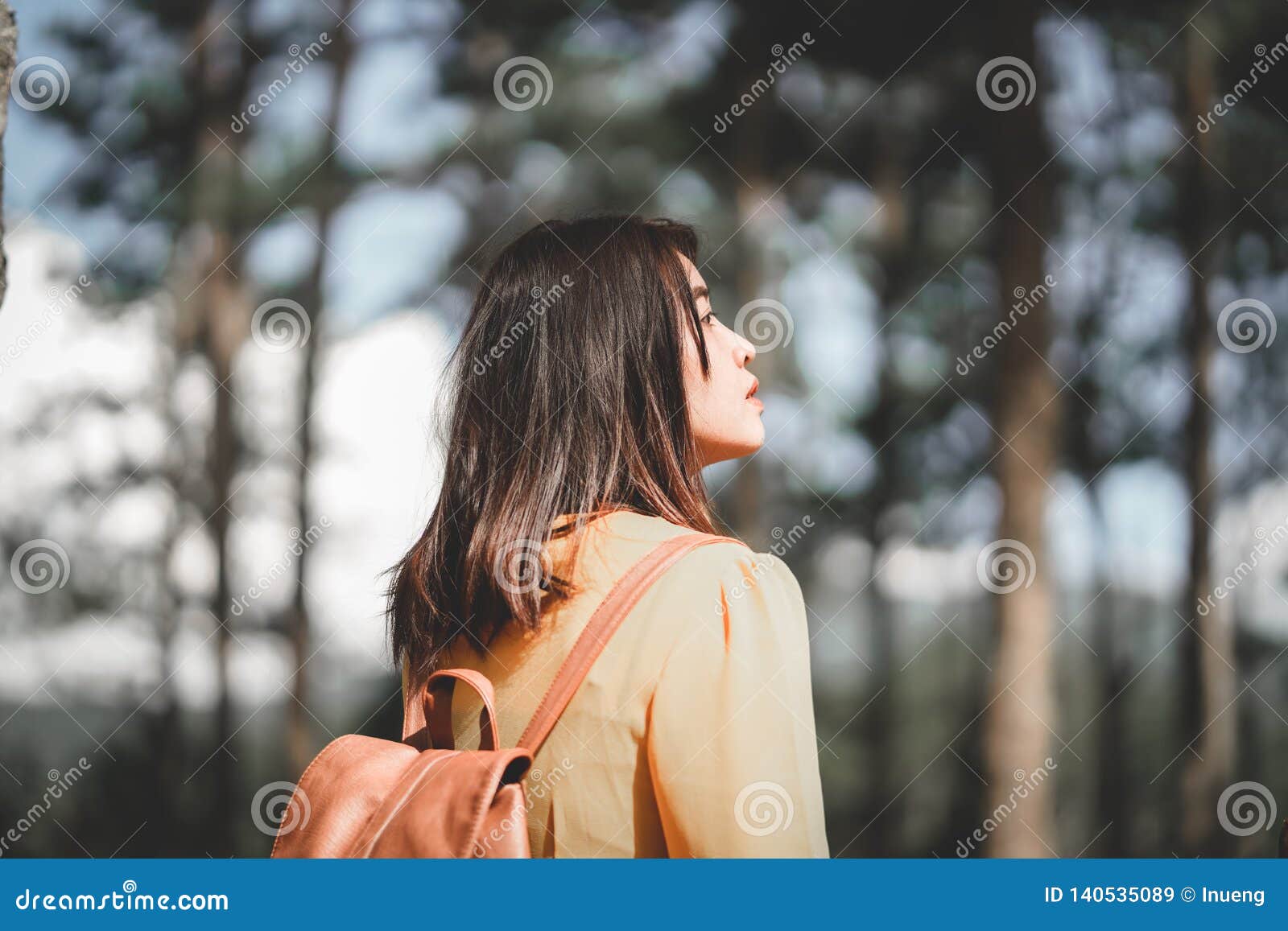 Asian girl traveler in yellow dress with backpack walking in the forest. Rear view.