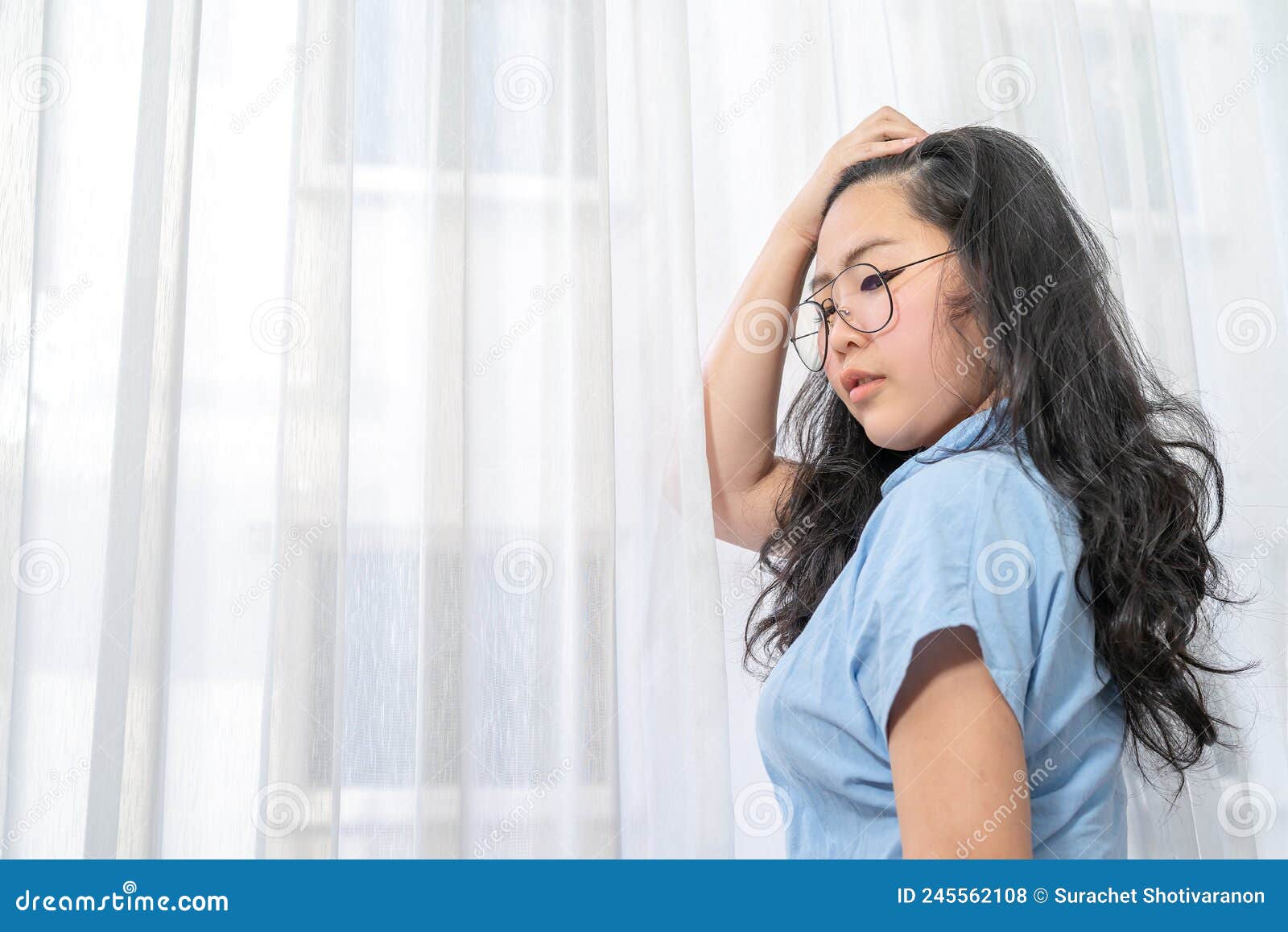 an asian glasses woman in blue shirt  poses by slicks her hair back in front of lightening curtain