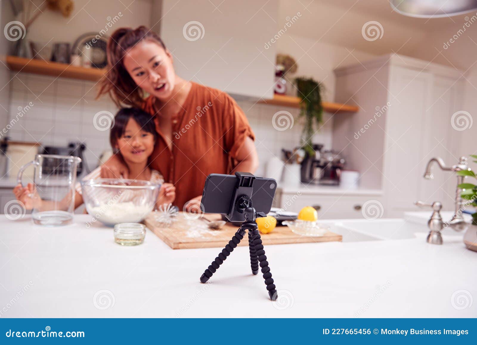 asian mother and daughter baking cupcakes in kitchen at home whilst on vlogging on mobile phone