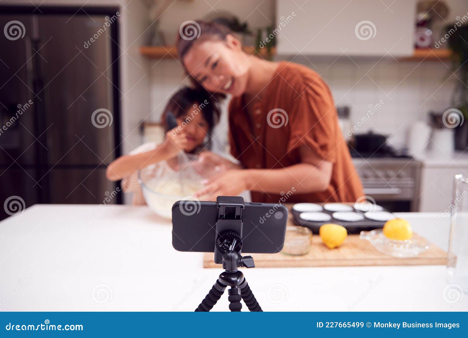 asian mother and daughter baking cupcakes in kitchen at home whilst on vlogging on mobile phone