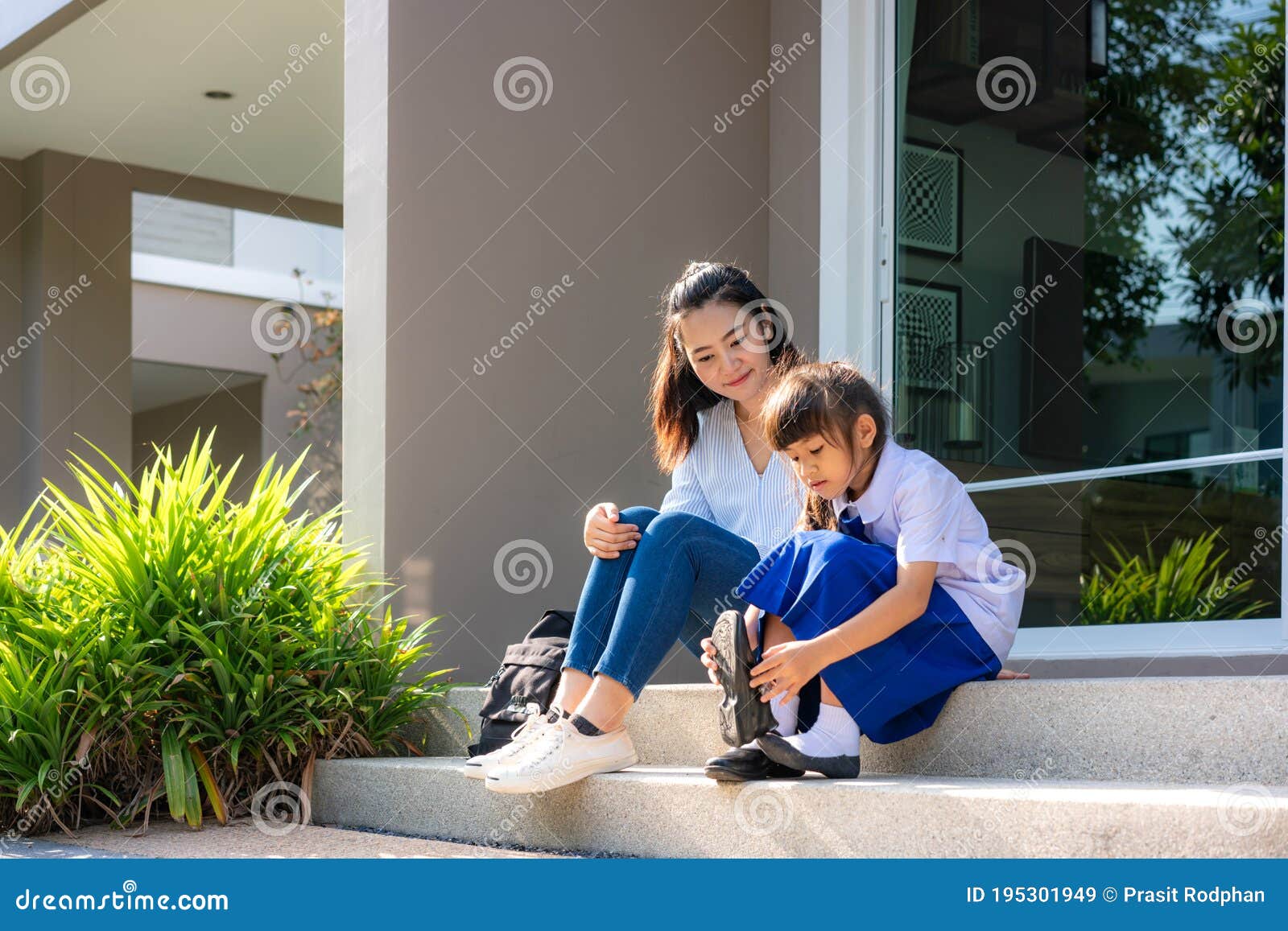 asian mother looking her daughter primary students in uniform to wearing their own shoes infront of home in the morning school