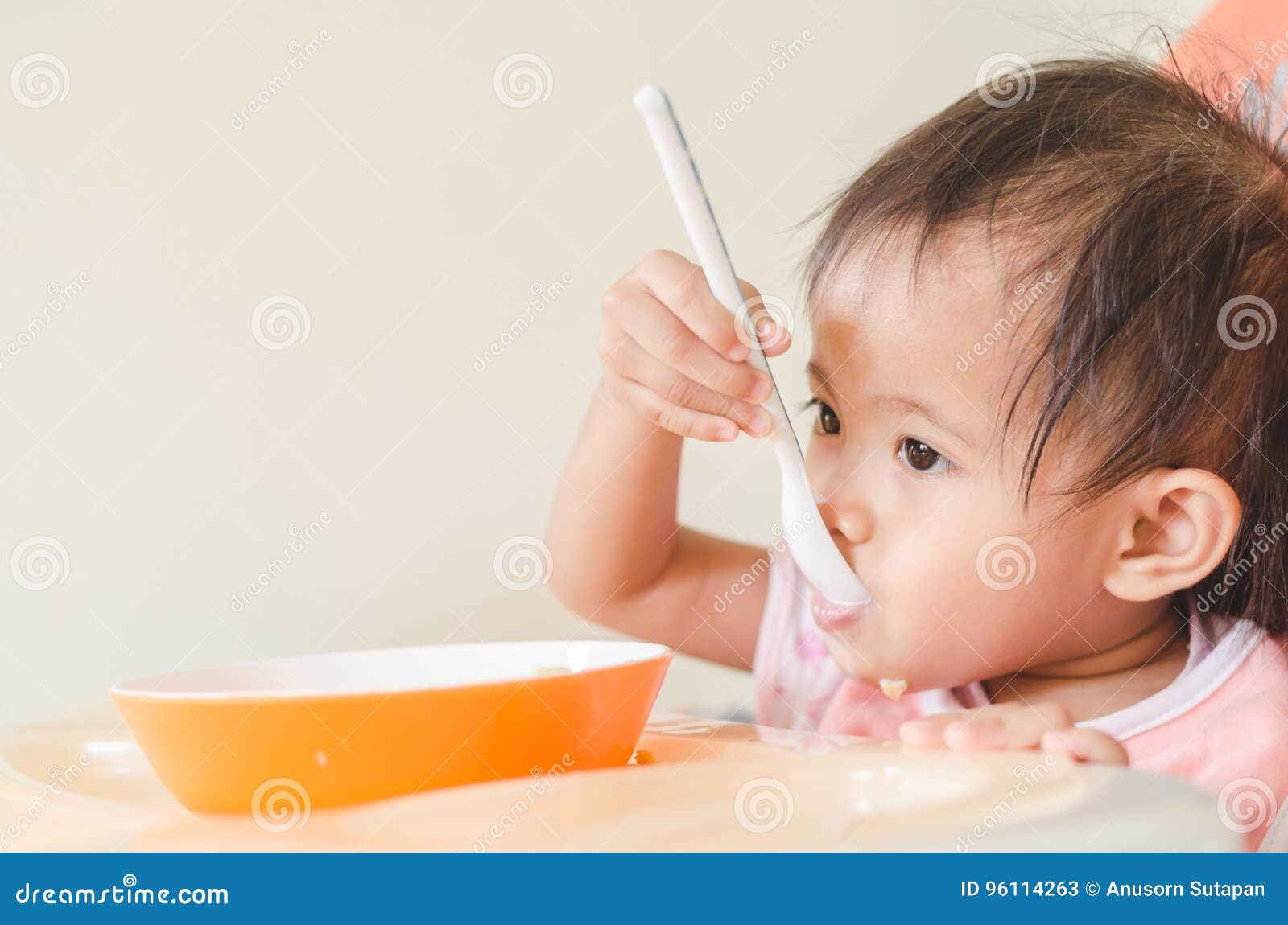 Asian Toddler Girl Eating Cereals With Milk On High Chair At Home