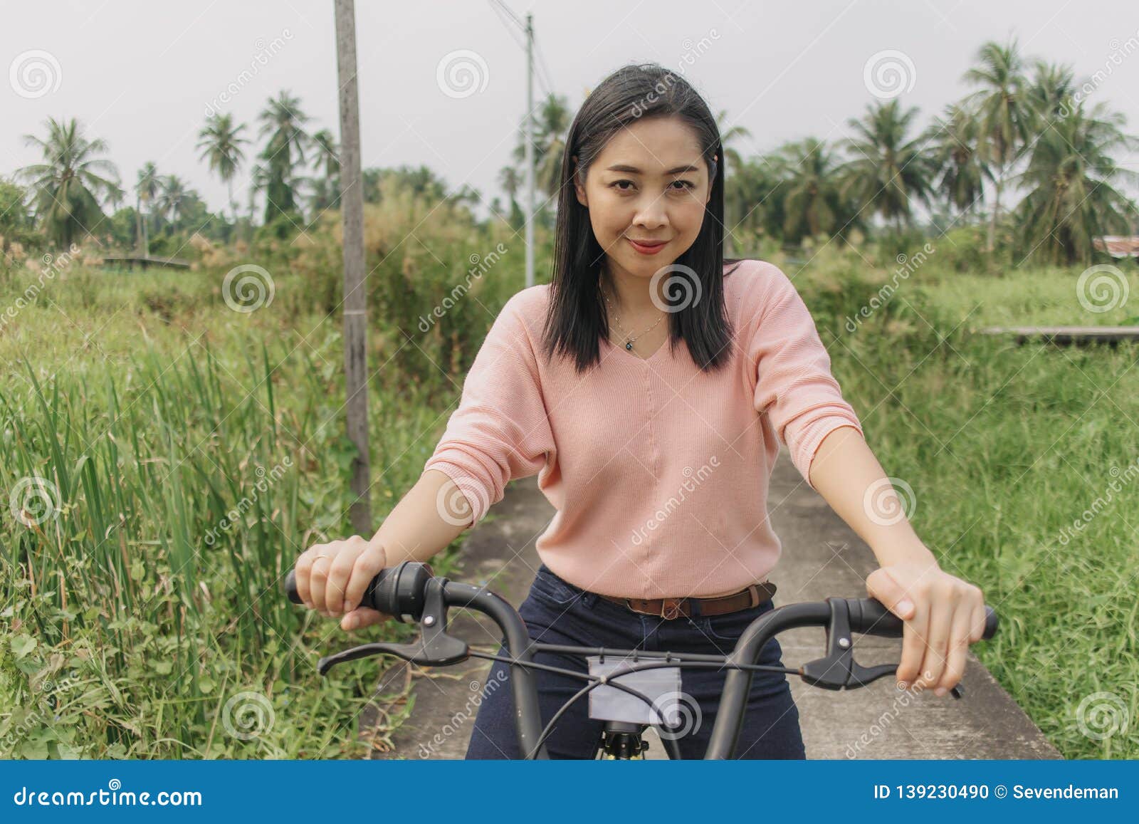Asian Woman Ride Bicycle On The Local Street With Greenery View Stock
