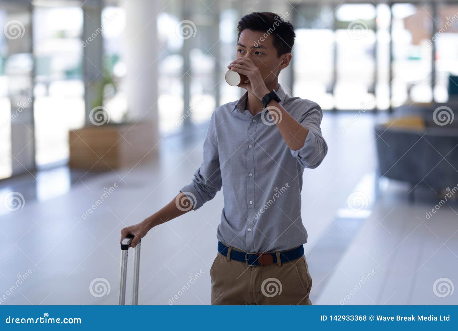 Asiatisk manlig ledare som har kaffe, medan gå med bagage i lobbyen. Främre sikt av den unga asiatiska manliga ledaren som har kaffe, medan gå med bagage i lobbyen på kontoret Han tänker