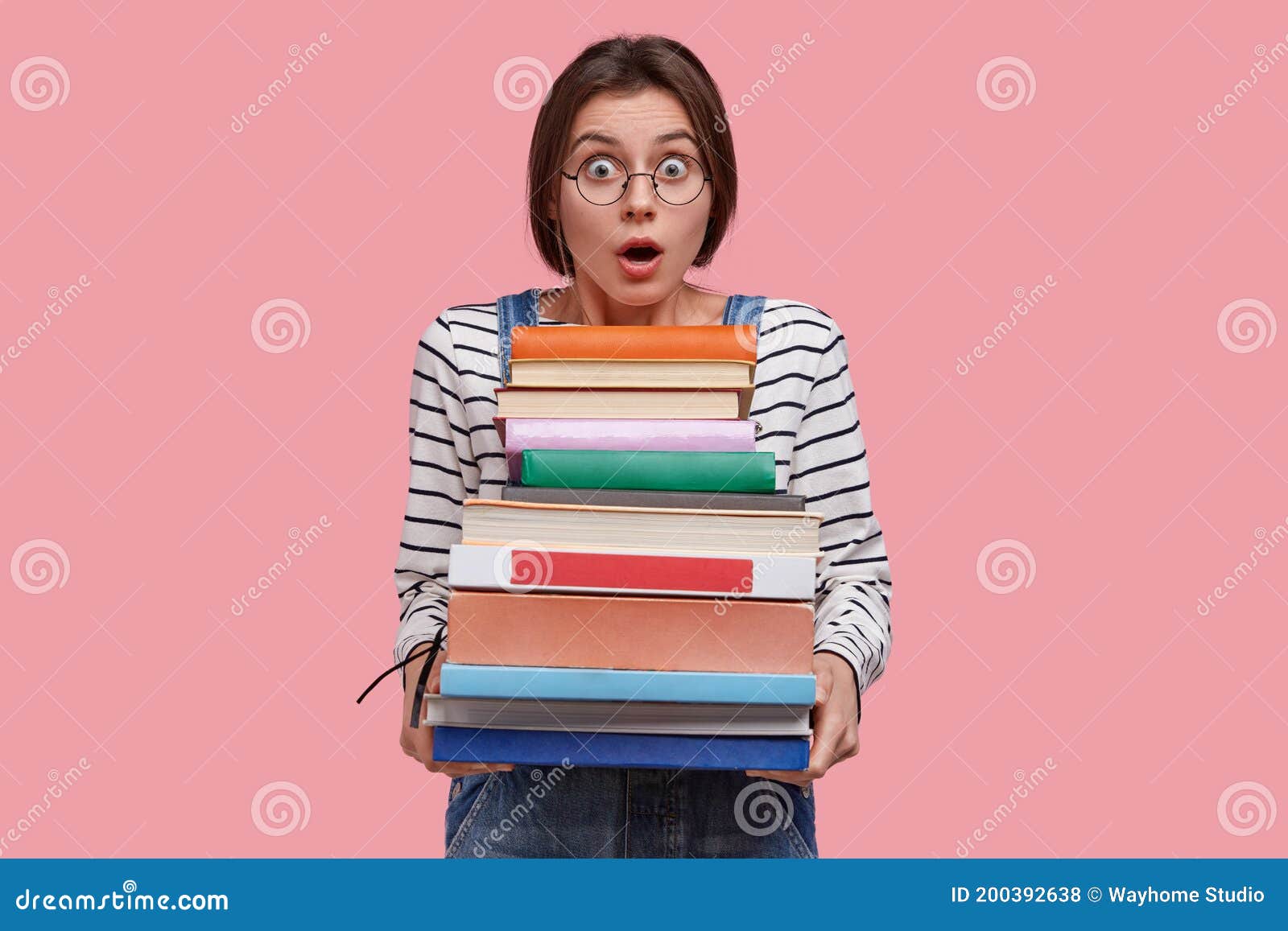 astounded young european woman with dark hair, dressed in striped clothes, carries many books, being stupefied to learn