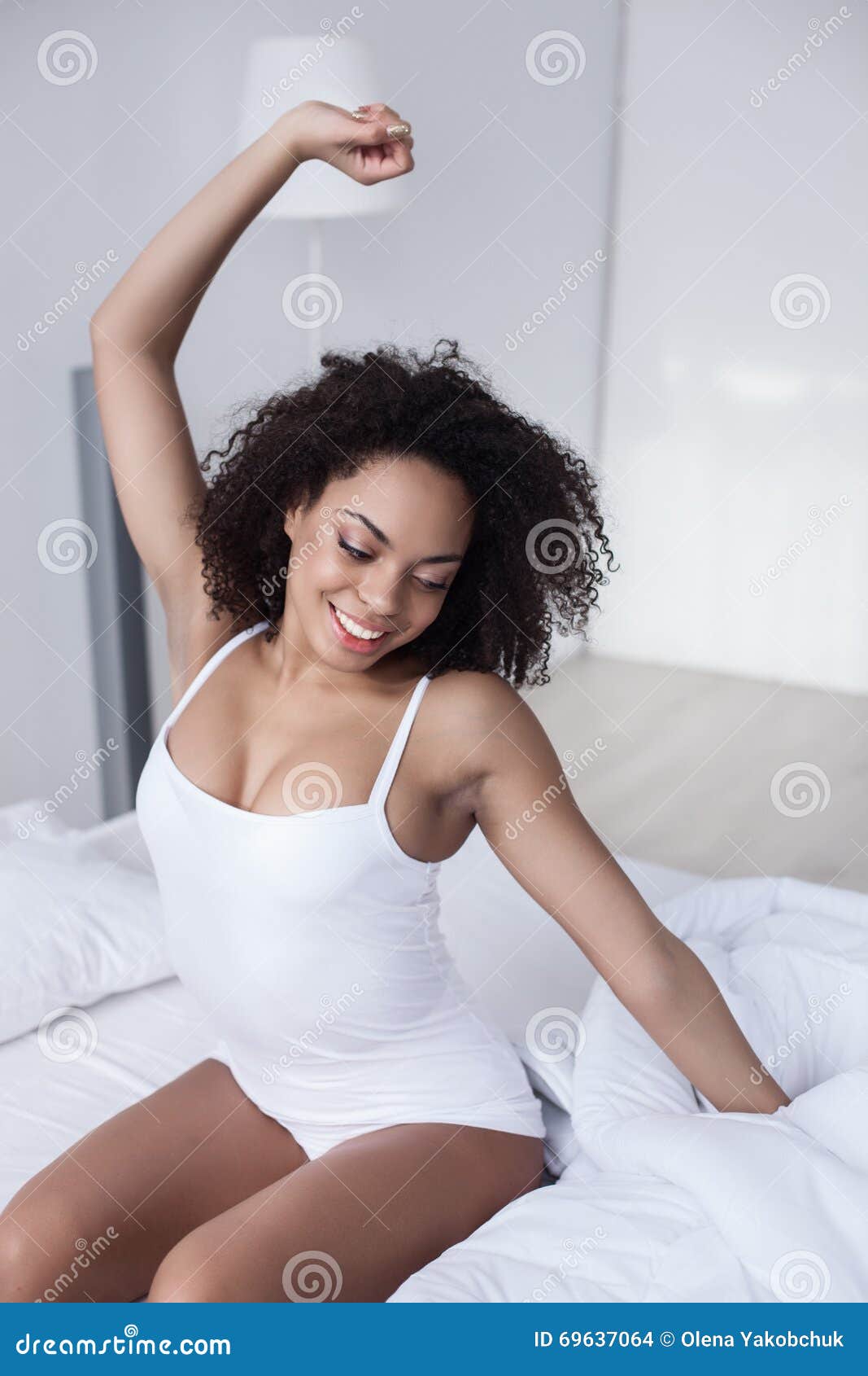 Attractive african woman stretching arms after sleeping. Beautiful young mulatto girl is waking up in the morning. She is sitting on bed with her hand outstretched. The lady is smiling happily